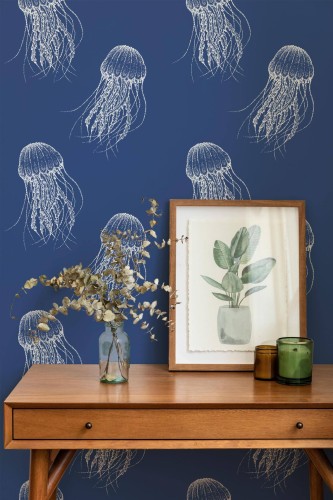 A navy blue jellyfish wallpaper features large white illustrated jellyfish with trailing tentacles in a repeating pattern, bringing a whimsical nautical touch to this serene interior. Styled in a cozy corner with mid-century modern wooden furniture and dried florals, the design suits coastal, bohemian, or kids’ room décors. This Fancy Walls wallpaper is available in peel and stick (self-adhesive polyester textile) or non-pasted (FSC mix-certified non-woven) materials, ideal for creative, ocean-inspired spaces.