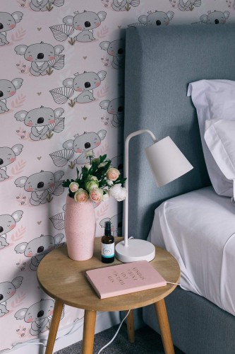 Cute Koala Wallpaper with soft pink background, showing whimsical koala illustrations and delicate leaf accents, styled in a cozy bedroom with a gray upholstered bed, wooden side table, pink vase of flowers, and a modern white lamp. The playful design adds a sweet, inviting charm to the space.