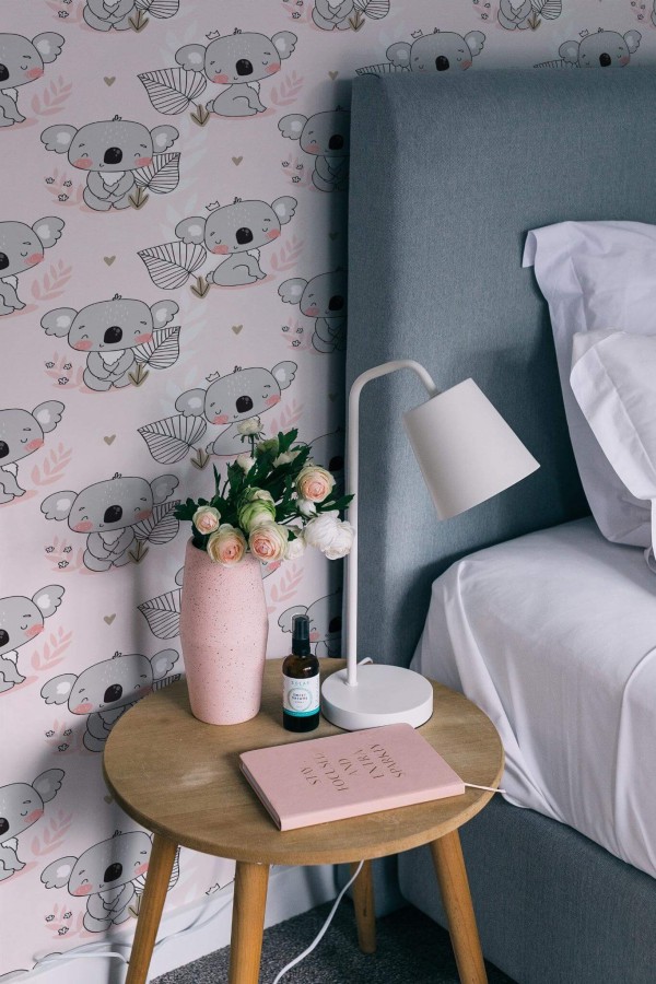 Cute Koala Wallpaper with soft pink background, showing whimsical koala illustrations and delicate leaf accents, styled in a cozy bedroom with a gray upholstered bed, wooden side table, pink vase of flowers, and a modern white lamp. The playful design adds a sweet, inviting charm to the space.