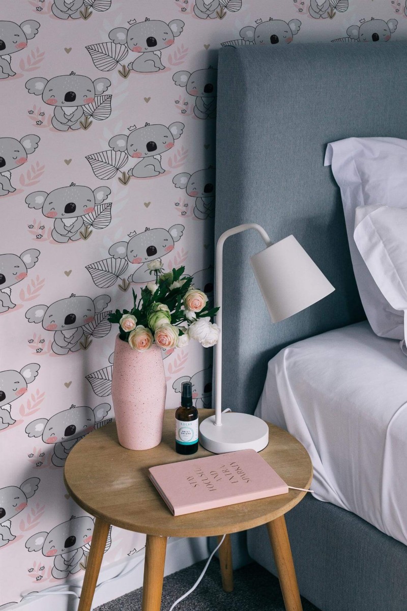 Cute Koala Wallpaper with soft pink background, showing whimsical koala illustrations and delicate leaf accents, styled in a cozy bedroom with a gray upholstered bed, wooden side table, pink vase of flowers, and a modern white lamp. The playful design adds a sweet, inviting charm to the space.
