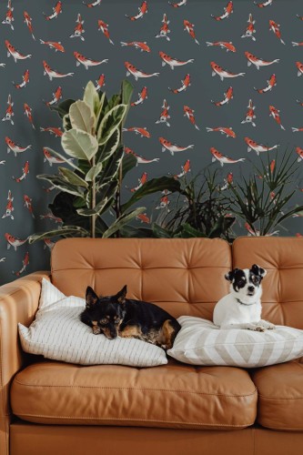 Living room decorated with dark gray koi fish wallpaper featuring orange-and-white koi swimming in a scattered pattern. A caramel-brown leather sofa sits in front, holding two small dogs—one black-and-tan and one white-and-black—relaxing on neutral striped pillows. Behind the sofa, tall green houseplants add natural texture, contrasting with the lively fish motif. The scene feels cozy yet dynamic, blending playful aquatic art with warm, modern interior décor.