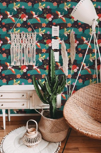 Maximalist Bird Wallpaper with vivid teal background, showcasing colorful birds, blooming flowers, and lively foliage, styled in a bohemian space with a macramé wall hanging, tall cactus, rattan chair, and a warm wood floor. The bold pattern infuses the room with an energetic, whimsical atmosphere.