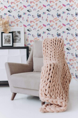 Modern nail salon wallpaper with colorful abstract illustrations of manicure tools, hands, polish bottles, and salon equipment on a light cream background. The novelty wallpaper features an isometric layout with soft pink, purple, and blue hues, ideal for beauty salons, powder rooms, or vanity corners. Pictured in a cozy setting with a neutral armchair, chunky knit throw blanket, and framed black-and-white portraits on a minimalist white cabinet, this peel and stick or non-pasted wallpaper design adds playful charm to professional or home décors.