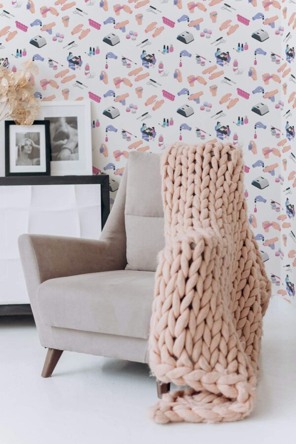 Modern nail salon wallpaper with colorful abstract illustrations of manicure tools, hands, polish bottles, and salon equipment on a light cream background. The novelty wallpaper features an isometric layout with soft pink, purple, and blue hues, ideal for beauty salons, powder rooms, or vanity corners. Pictured in a cozy setting with a neutral armchair, chunky knit throw blanket, and framed black-and-white portraits on a minimalist white cabinet, this peel and stick or non-pasted wallpaper design adds playful charm to professional or home décors.