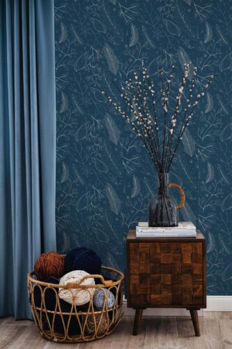 A cozy interior with Fancy Walls peel and stick or non-pasted wallpaper featuring a navy blue winter botanical design. The wall showcases hand-drawn winter foliage, berries, and pine leaves in soft gray outlines against a deep navy background. A wooden mid-century side table with a glass vase and books, along with a woven basket filled with yarn, adds warmth and texture to the scene. The Fancy Walls peel and stick or non-pasted wallpaper brings seasonal charm to this elegant living space.