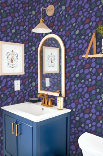 A bold, eclectic powder room featuring Fancy Walls “Blue Wildcat Brush” wallpaper, designed with an abstract animal print in deep navy, indigo, sage green, and wine red brushstroke spots. The playful, stylized wildcat pattern adds dynamic movement and texture to the walls, creating a modern, statement-making backdrop. The wallpaper pairs beautifully with a navy blue vanity, brass faucet, arched wood-framed mirror, and light wood accents. Available as peel and stick or non-pasted wallpaper, this eco-conscious design is PVC-free and printed with GreenGuard Gold certified inks for vivid color and safe indoor air quality.