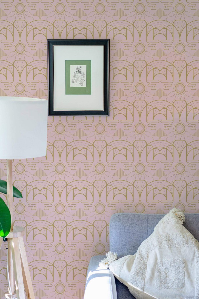 A soft, pastel pink wall adorned with an elegant geometric Art Deco design featuring golden fan-like shapes and intricate lines. The design creates a sense of luxury and sophistication, making it a perfect statement piece for a modern living room or bedroom. A white lamp and a cozy gray sofa with a fluffy pillow complement the vibrant and stylish wall decor. The geometric pattern adds a bold touch, while the soft hues of the wallpaper balance the overall aesthetic. The Fancy Walls peel and stick or non-pasted wallpaper transforms your space with timeless Art Deco charm.