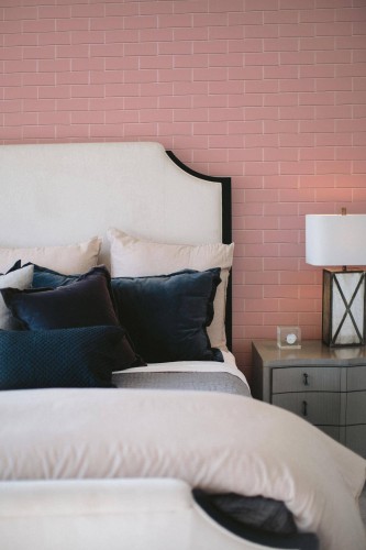 A cozy bedroom interior featuring Fancy Walls pink brick wallpaper in a classic horizontal layout. The wallpaper showcases soft, matte pink bricks with subtle grout lines, creating a gentle geometric rhythm that adds structure without overwhelming the space. Styled in a modern eclectic setting, the room includes a cream upholstered headboard, navy velvet accent pillows, and a light wood nightstand with a cross-frame lamp. The wallpaper is available in two eco-conscious formats: peel and stick (self-adhesive polyester textile) or non-pasted (FSC mix-certified non-woven), both printed with GreenGuard Gold certified inks for vibrant color and safe indoor air quality.