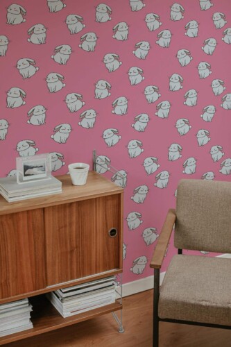 Pink bunny wallpaper featuring multiple white rabbits on a bold pink background, displayed in a cozy corner with a mid-century wooden sideboard, neutral armchair, and minimal decor.