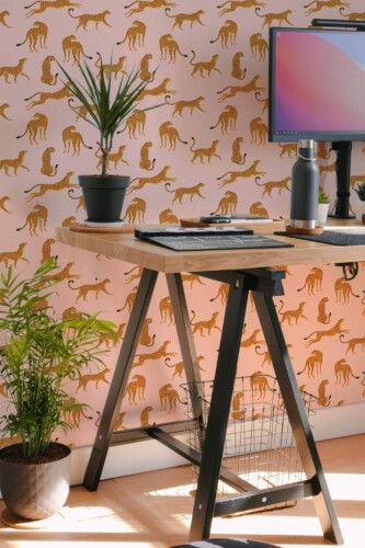 Pink Cheetah Wallpaper featuring stylized gold cheetah silhouettes on a soft pink background, styled in a modern home office with a wooden desk, black metal legs, potted plants, and a desktop monitor. The eclectic animal print adds bold personality and a playful accent to the workspace.