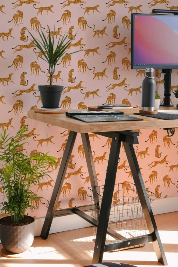 Pink Cheetah Wallpaper featuring stylized gold cheetah silhouettes on a soft pink background, styled in a modern home office with a wooden desk, black metal legs, potted plants, and a desktop monitor. The eclectic animal print adds bold personality and a playful accent to the workspace.