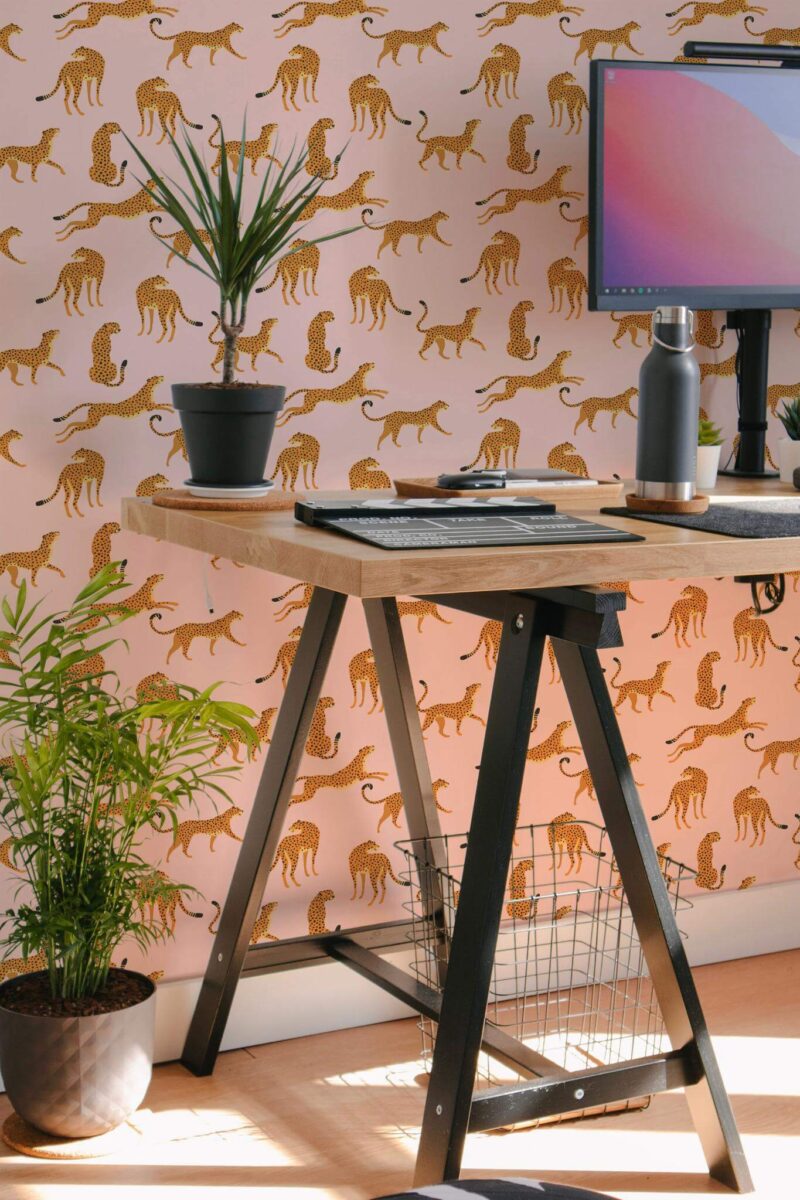 Pink Cheetah Wallpaper featuring stylized gold cheetah silhouettes on a soft pink background, styled in a modern home office with a wooden desk, black metal legs, potted plants, and a desktop monitor. The eclectic animal print adds bold personality and a playful accent to the workspace.