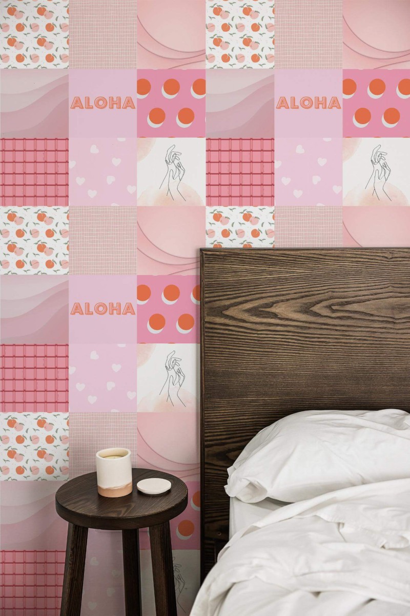 Bedroom featuring a whimsical pink collage wallpaper composed of square tiles with varied patterns, including hearts, fruits, “Aloha” text, and minimalist line-drawn figures. The patchwork design layers soft pastel hues, geometric shapes, and playful prints to create a vibrant accent wall. A dark wooden headboard anchors the right side, complemented by crisp white bedding. A small round wooden nightstand with a simple white candle stands nearby, adding a cozy, modern contrast.
