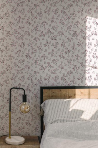Close-up of a cozy bedroom featuring ditsy floral wallpaper with hand-drawn branches and small red blossoms on a warm gray background. The botanical pattern creates a soft, vintage-inspired look ideal for minimalist or modern rustic interiors. A wood-accented bed and marble lamp base add texture and contrast to the soft wallpaper design.