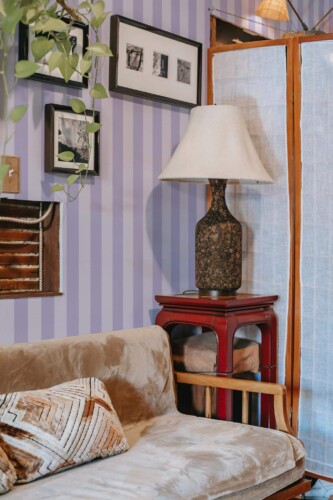 Pastel Purple Stripes Wallpaper with subtle lilac vertical bands, styled in a cozy living area featuring a velvet sofa, red accent table, rustic lamp, framed photos, and hanging greenery. The soothing striped pattern adds modern elegance and a gentle pop of color to the space.