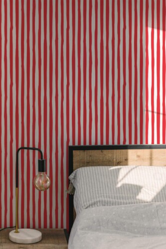 Bedroom accent wall displaying bold red stripes wallpaper in a hand-drawn style. The vertical stripes create a lively, graphic effect against the neutral background. A wood-framed bed sits on the right, dressed in soft gray-striped linens, while a modern black-and-brass lamp with a round exposed bulb stands on the left. Warm daylight filters into the space, enhancing the contrast of the vibrant red stripes and highlighting the minimal yet striking décor.