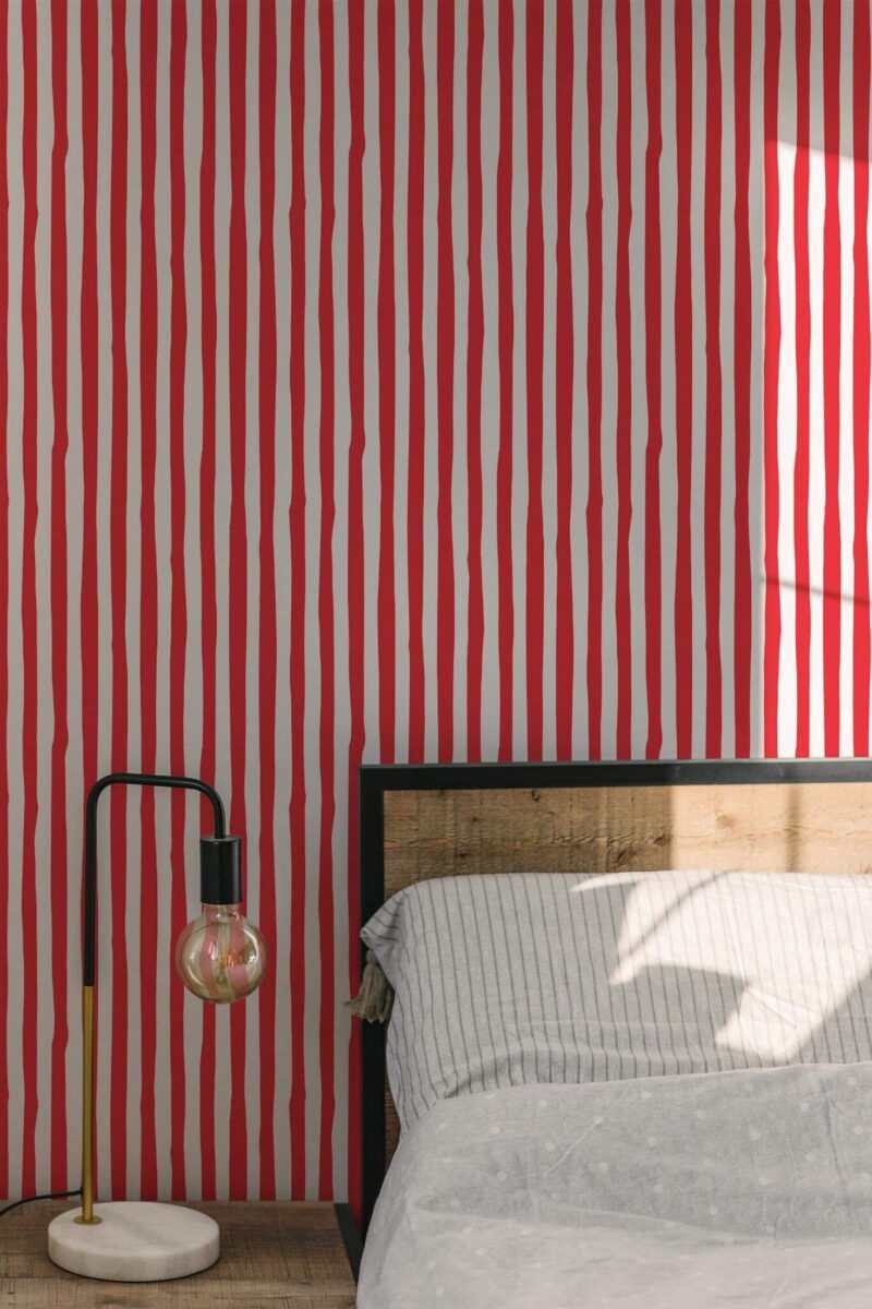 Bedroom accent wall displaying bold red stripes wallpaper in a hand-drawn style. The vertical stripes create a lively, graphic effect against the neutral background. A wood-framed bed sits on the right, dressed in soft gray-striped linens, while a modern black-and-brass lamp with a round exposed bulb stands on the left. Warm daylight filters into the space, enhancing the contrast of the vibrant red stripes and highlighting the minimal yet striking décor.