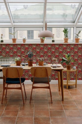 Mid-century style dining room with terracotta floor tiles and a glass ceiling features a bold accent wall covered in Fancy Walls' "Red Bold Mushrooms wallpaper." The wallpaper design showcases clusters of large, whimsical mushrooms in cream, taupe, and forest green, intricately illustrated with stylized caps and stems. The mushrooms are repeated in a tight, symmetrical pattern against a rich, brick-red background, creating a dramatic yet earthy visual impact. Paired with minimalist wood furniture and potted plants on the window ledge, the space exudes a warm, boho-modern charm. The wallpaper’s vintage-inspired botanical motif harmonizes with the natural light and greenery, making it ideal for kitchens, sunrooms, or creative studios looking for a vibrant and organic statement feature.