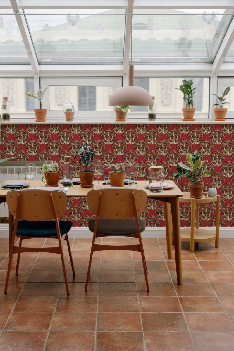 Mid-century style dining room with terracotta floor tiles and a glass ceiling features a bold accent wall covered in Fancy Walls' "Red Bold Mushrooms wallpaper." The wallpaper design showcases clusters of large, whimsical mushrooms in cream, taupe, and forest green, intricately illustrated with stylized caps and stems. The mushrooms are repeated in a tight, symmetrical pattern against a rich, brick-red background, creating a dramatic yet earthy visual impact. Paired with minimalist wood furniture and potted plants on the window ledge, the space exudes a warm, boho-modern charm. The wallpaper’s vintage-inspired botanical motif harmonizes with the natural light and greenery, making it ideal for kitchens, sunrooms, or creative studios looking for a vibrant and organic statement feature.