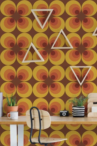 Retro 70s circle wallpaper in warm orange, yellow, and brown tones, displayed in a creative workspace with a wooden desk, triangular wall shelves, potted succulents, and a modern chair for a vibrant, nostalgic flair.