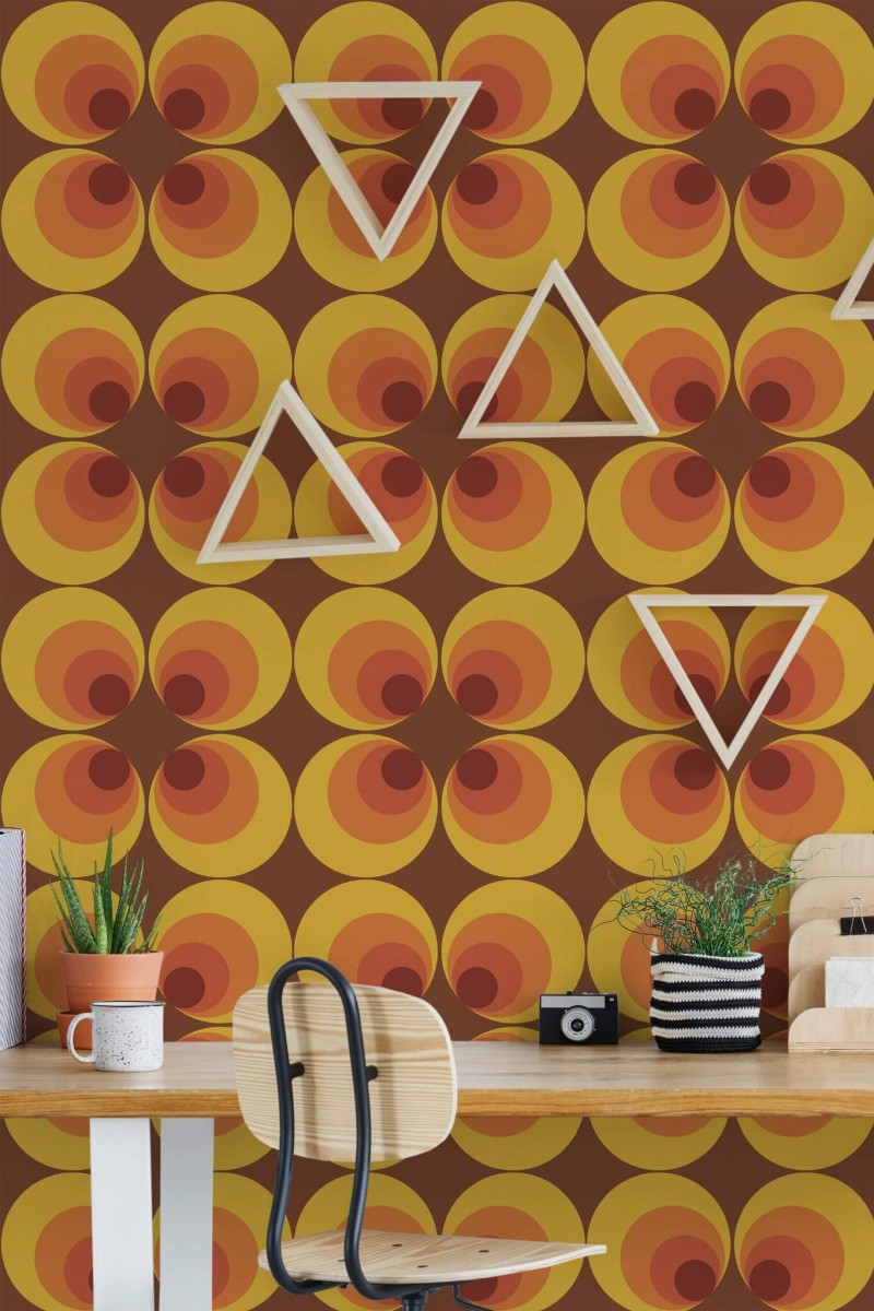 Retro 70s circle wallpaper in warm orange, yellow, and brown tones, displayed in a creative workspace with a wooden desk, triangular wall shelves, potted succulents, and a modern chair for a vibrant, nostalgic flair.