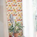 Retro Psychedelic Wallpaper featuring playful mushroom and floral motifs in warm oranges, yellows, and pinks on a soft pastel background, styled in a bright interior with sheer curtains, marble flooring, a modern standing fan, and a potted plant. The bold, vintage-inspired pattern adds a lively, bohemian accent to the room.
