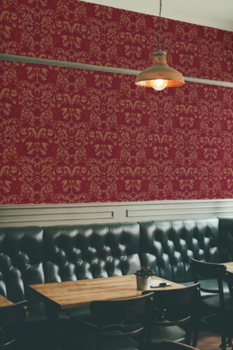 Burgundy Elegance Wallpaper with an ornate ochre damask pattern on a rich red background, styled in a sophisticated bar interior featuring tufted black leather seating, wooden tables, and a warm industrial pendant lamp. The luxurious design adds timeless charm and a refined atmosphere to the space.