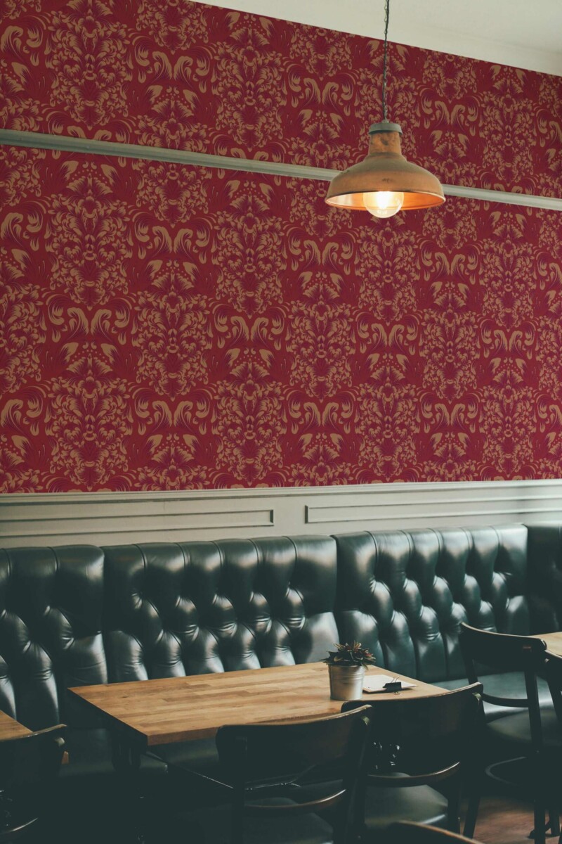 Burgundy Elegance Wallpaper with an ornate ochre damask pattern on a rich red background, styled in a sophisticated bar interior featuring tufted black leather seating, wooden tables, and a warm industrial pendant lamp. The luxurious design adds timeless charm and a refined atmosphere to the space.