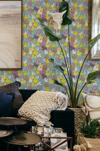 A cozy, eclectic living room with a feature wall covered in multicolored rubber duck illustrations. The playful duck pattern includes yellow, pink, brown, mint, and lavender tones on a soft teal background, creating a fun and vibrant vibe. Styled with woven blankets, tropical houseplants, and modern furnishings, this space balances charm and sophistication. The wall is covered in Fancy Walls peel and stick or non-pasted wallpaper, offering a PVC-free, eco-friendly option perfect for creative accent walls.