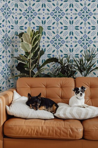Scandinavian Folk Wallpaper with symmetrical blue tile-inspired motifs on a white background, styled behind a caramel leather sofa where two small dogs rest on cushions, surrounded by lush indoor plants. The playful Nordic pattern adds cozy charm and a modern twist to this inviting living room setting.