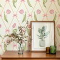 Beige Bow Blooms wallpaper featuring pink tulips and bows arranged in a diamond pattern. Displayed in a warm, minimal interior with a wooden console table, framed botanical art, and dried foliage accents.