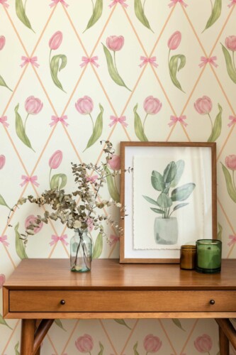 Beige Bow Blooms wallpaper featuring pink tulips and bows arranged in a diamond pattern. Displayed in a warm, minimal interior with a wooden console table, framed botanical art, and dried foliage accents.