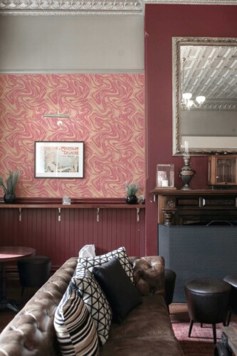 An elegant sitting room featuring Fancy Walls peel and stick or non-pasted wallpaper in a pink and cream marble swirl pattern. The wallpaper showcases flowing, abstract curves in warm blush tones layered over a soft beige background, creating a dynamic yet refined visual texture. This vintage-inspired space includes a tufted leather sofa with patterned cushions, a carved wooden mantel, and ornate ceiling moldings, all complemented by deep burgundy wall accents and antique décor. The stylish mix of old-world charm and modern artistic wallpaper results in a cozy yet sophisticated lounge atmosphere. The wallpaper is PVC-free, available in self-adhesive polyester textile or FSC mix-certified non-woven, and printed with GreenGuard Gold certified inks for vivid, safe, and eco-conscious indoor décor.