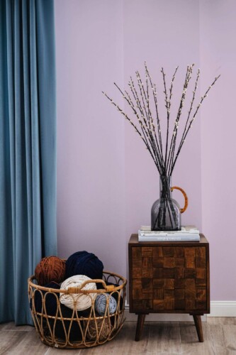Pastel Aesthetic Lilac Solid Color Wallpaper with a soft, dusty lilac hue, styled in a cozy space with a teal curtain, a dark wooden side table holding a glass vase of branches, and a woven basket filled with colorful yarn. The minimalist design and gentle color palette create a calm, modern ambiance.