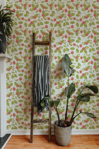Interior space decorated with strawberry summer wallpaper featuring bright pink strawberries and leafy green foliage on a white backdrop. A rustic wooden ladder leans against the accent wall, draped with a striped gray-and-black textile. In front, a large potted tropical plant with broad, dark-green leaves sits in a woven basket, adding a natural, bohemian element. The light wood floor and subtle fireplace edge at left complete this fresh, vibrant setting that mixes rustic charm with playful botanical style.