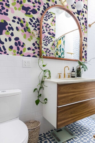 A bold and cheerful bathroom interior featuring Fancy Walls “Playful Spotscape” wallpaper, styled with abstract animal print spots in lilac pink, lime green, and deep navy on a soft cream background. The wallpaper creates a lively, artistic accent above classic white subway tiles. A wooden vanity with brass fixtures, a curved wood-framed mirror, leafy green plants, and patterned floor tiles complete the modern eclectic look. This design is available as peel and stick or non-pasted wallpaper, printed with eco-friendly GreenGuard Gold certified inks on PVC-free materials—perfect for vibrant, personality-filled spaces.