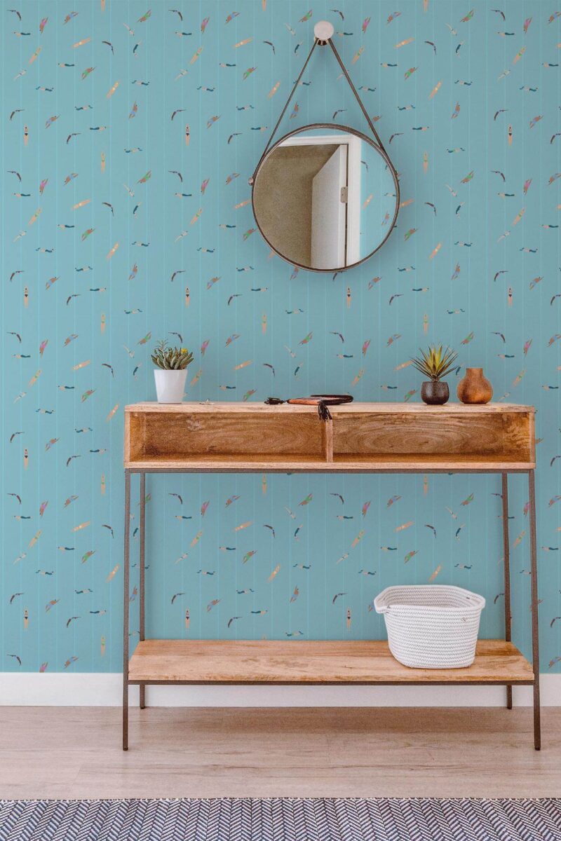 Turquoise wallpaper with playful swimmer figures depicting a lively pool scene, featuring intricate design details. Shown in a modern entryway with a rustic wooden console, a round mirror, and potted succulents, creating a chic, airy atmosphere.