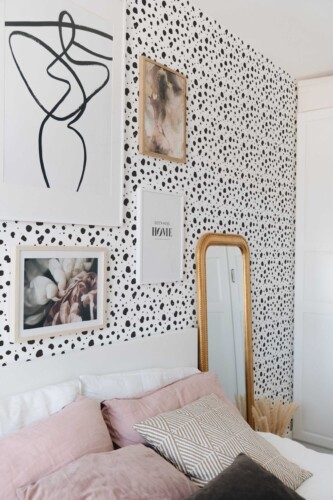 Bedroom accent wall decorated with tiny monochrome cheetah print wallpaper in black and white. A collection of framed art, including abstract line drawings and floral photography, hangs on the wall above a white headboard. Soft pink and neutral-toned pillows layer across the bed, creating a calm, modern vibe. A tall gold-framed mirror leans against the wall, reflecting the printed pattern and adding warmth. Overall, the scene feels airy, stylish, and playful.