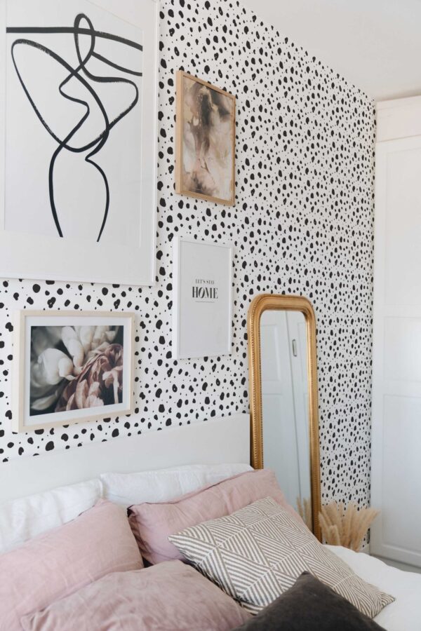 Bedroom accent wall decorated with tiny monochrome cheetah print wallpaper in black and white. A collection of framed art, including abstract line drawings and floral photography, hangs on the wall above a white headboard. Soft pink and neutral-toned pillows layer across the bed, creating a calm, modern vibe. A tall gold-framed mirror leans against the wall, reflecting the printed pattern and adding warmth. Overall, the scene feels airy, stylish, and playful.