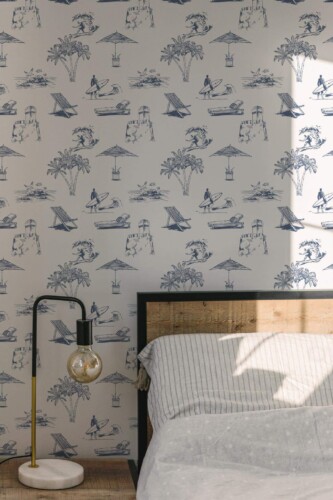 Toile Beach Wallpaper with vintage-style blue coastal scenes on a soft white background, styled in a calm bedroom featuring a wooden bed frame, neutral bedding, and a modern glass bulb lamp. The coastal motifs include beach umbrellas, surfers, and palm trees, adding relaxed seaside charm to the space.