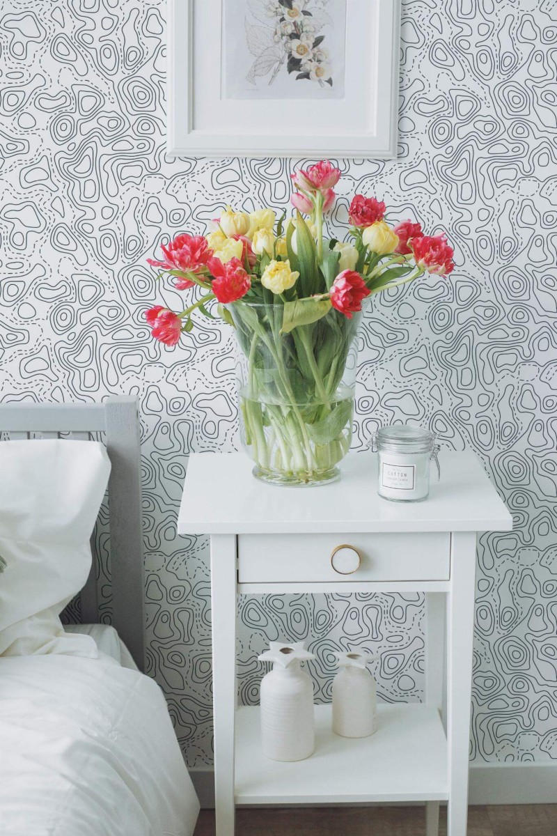Topographic Wallpaper with black abstract contour lines on a white background, styled in a bright bedroom with a white nightstand, colorful tulips in a glass vase, and minimal decor. The modern line art pattern adds subtle movement and visual interest to the tranquil space.