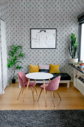 Compact dining area showcasing black and white bamboo-like striped wallpaper as a dramatic accent wall. A small round white table with wooden legs sits at the center, flanked by two pink upholstered chairs. Behind it, a low bench adorned with yellow and gray throw pillows offers additional seating. Several potted plants—such as a tall cactus and lush tropical greenery—surround the space, adding a fresh touch. Minimalist artwork in a black frame hangs above the bench, completing the bright, modern look.