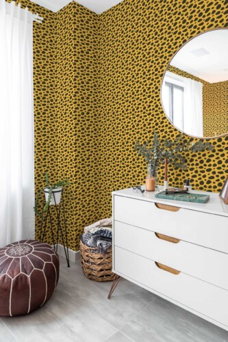 Bright corner of a modern room featuring vibrant cheetah print wallpaper in deep yellow and black, wrapping around two adjoining walls. A sleek white dresser with mid-century legs stands on the right, topped by a round mirror that reflects natural light. On the left, tall white curtains and a brown leather pouf add warmth. A woven basket filled with cozy blankets sits nearby, while dried greenery in a vase brings a subtle organic touch. The gray tile flooring completes the fresh, eclectic setting.