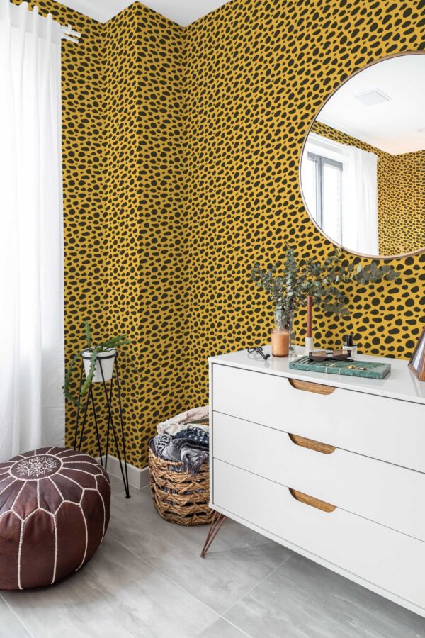 Bright corner of a modern room featuring vibrant cheetah print wallpaper in deep yellow and black, wrapping around two adjoining walls. A sleek white dresser with mid-century legs stands on the right, topped by a round mirror that reflects natural light. On the left, tall white curtains and a brown leather pouf add warmth. A woven basket filled with cozy blankets sits nearby, while dried greenery in a vase brings a subtle organic touch. The gray tile flooring completes the fresh, eclectic setting.