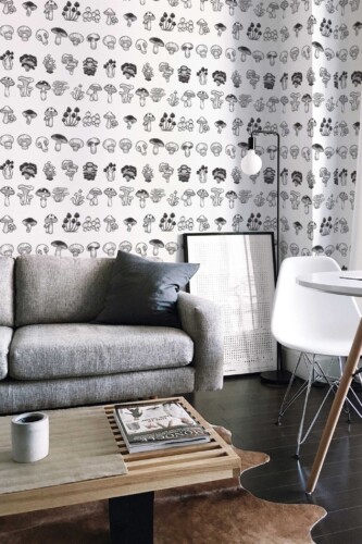 A modern living room showcasing the Handdrawn Mushroom Magic wallpaper, featuring delicate, monochrome, hand-drawn mushroom illustrations on a clean white background. The design brings an artistic botanical accent to the space, complementing a minimalist interior style. In the room, a sleek grey sofa pairs with a minimalist white accent chair, and a wooden coffee table adds warmth to the dark hardwood flooring. Framed artwork and subtle decor details enhance the contemporary feel, creating a refined, nature-inspired ambiance that blends modern elegance with organic charm