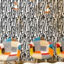 Eclectic interior featuring a bold black and white abstract wallpaper with hand-drawn tall mushrooms and wavy stems, arranged in a dense vertical pattern. The wallpaper creates a high-contrast graphic backdrop in a modern lounge space, paired with colorful patchwork armchairs in mid-century style. A white sculptural coffee table and trio of industrial copper pendant lights add warmth and balance to the vivid, artistic atmosphere. Ideal for creative spaces, cafés, or accent walls.