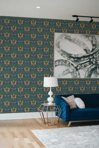 Living room with an accent wall covered in a dark blue wallpaper featuring art deco-style palm motifs and stylized wildcats in gold and green tones. A navy blue velvet sofa rests on the right, paired with a gold geometric side table topped by a white lamp. A large abstract canvas in gray and white hangs above the couch, contrasting the vibrant pattern. The hardwood floor and minimal décor elements complete the bright, sophisticated setting.