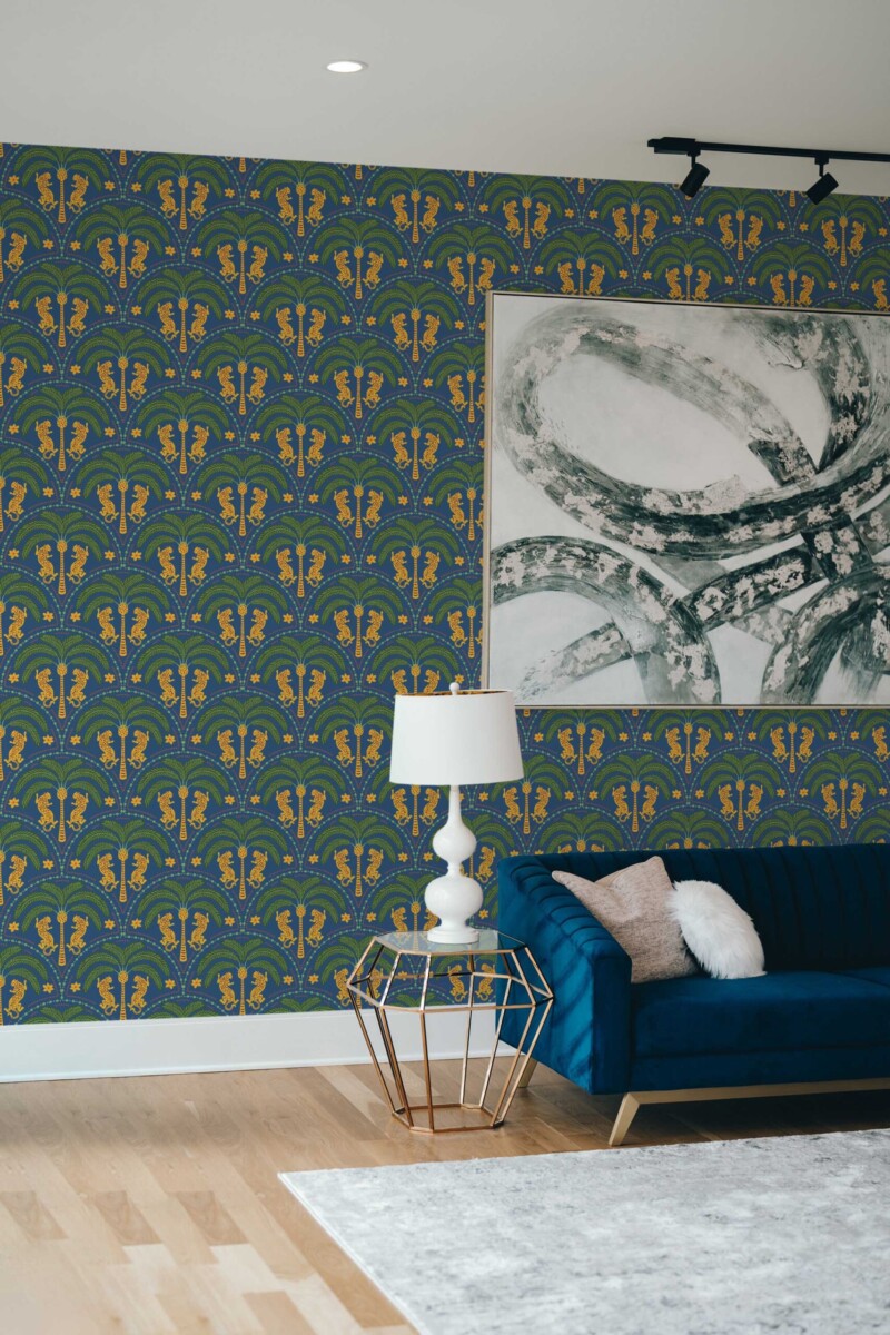 Living room with an accent wall covered in a dark blue wallpaper featuring art deco-style palm motifs and stylized wildcats in gold and green tones. A navy blue velvet sofa rests on the right, paired with a gold geometric side table topped by a white lamp. A large abstract canvas in gray and white hangs above the couch, contrasting the vibrant pattern. The hardwood floor and minimal décor elements complete the bright, sophisticated setting.