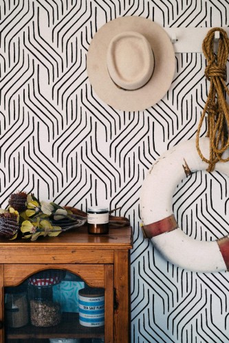 Seamless Modern Brushstroke wallpaper with bold black brush lines forming a dynamic, abstract pattern on a white background, styled in a coastal-inspired nook featuring a rustic wooden cabinet, a hanging lifebuoy, rope accents, and a neutral-toned hat. The striking black-and-white design adds a modern, artistic flair to the space.