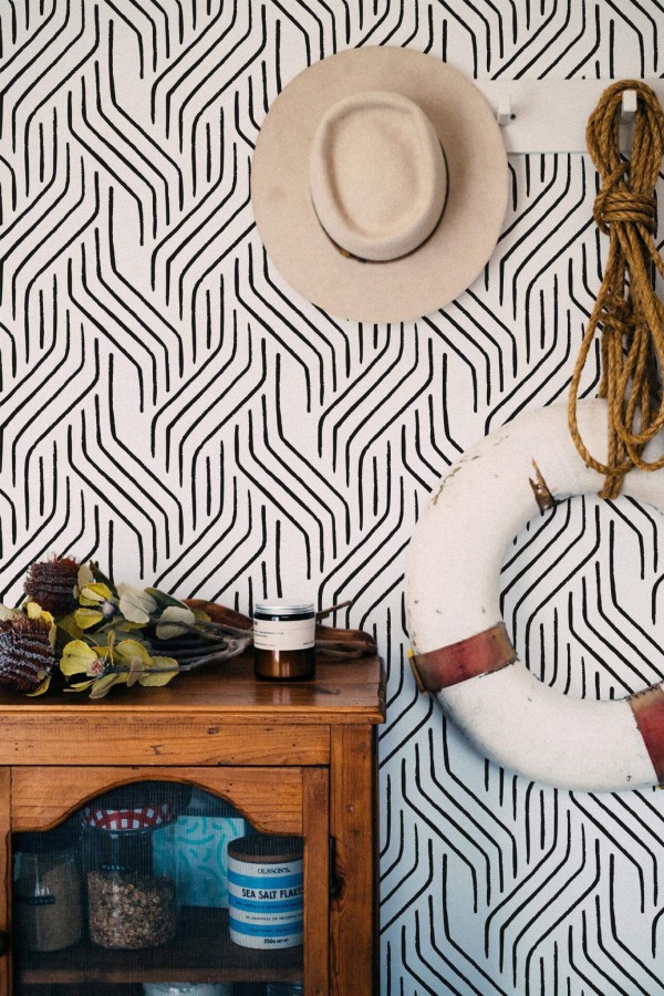 Seamless Modern Brushstroke wallpaper with bold black brush lines forming a dynamic, abstract pattern on a white background, styled in a coastal-inspired nook featuring a rustic wooden cabinet, a hanging lifebuoy, rope accents, and a neutral-toned hat. The striking black-and-white design adds a modern, artistic flair to the space.